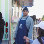 Man in blue apron and beanie standing outdoors near a food truck.