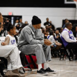 A man in gray sportswear sits courtside at a basketball game.