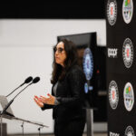 Woman speaking at a podium during a formal event with multiple microphones.