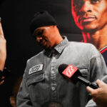 A man in a beanie and gray shirt speaks to reporters with microphones.