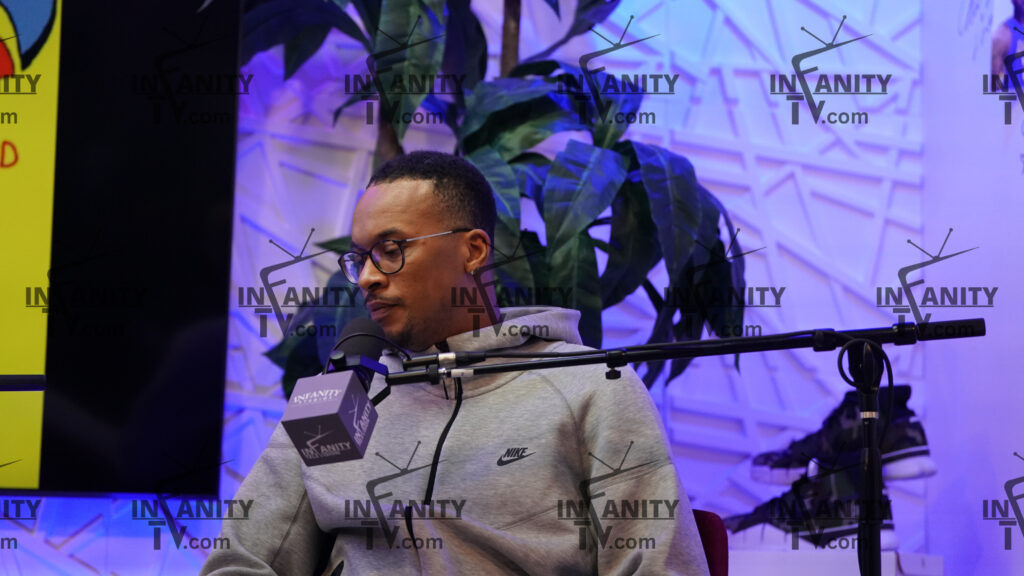 Man wearing glasses and a gray hoodie, sitting indoors with plants in the background.