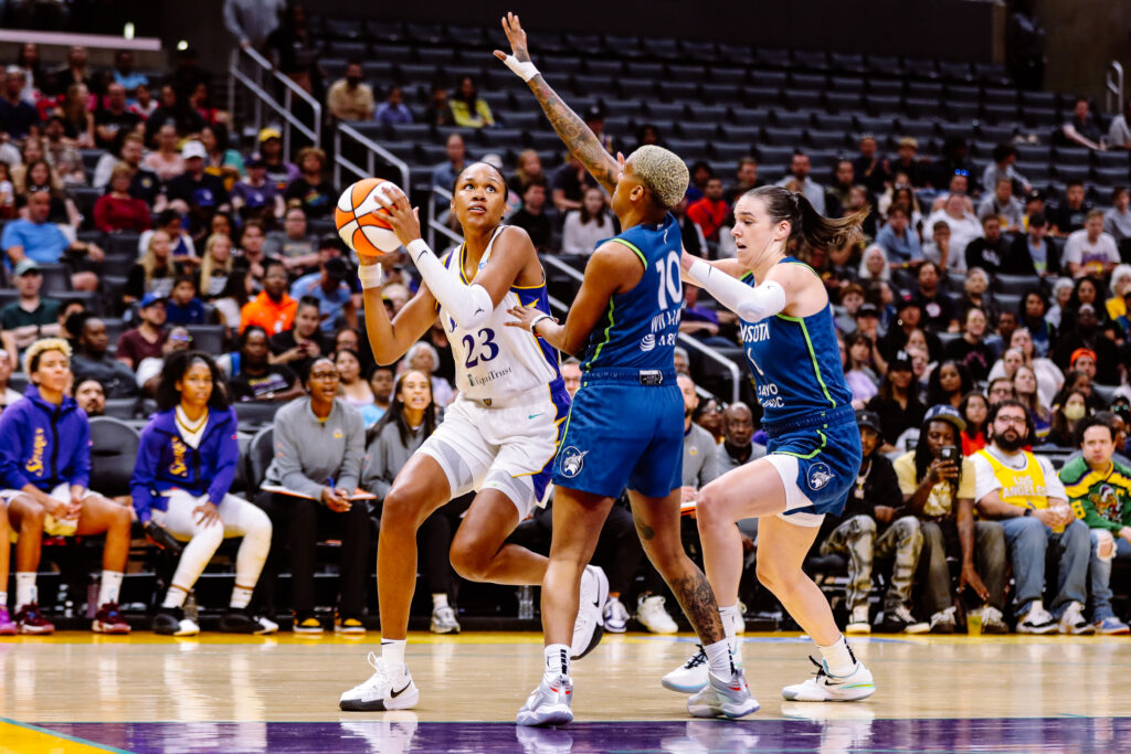 Female basketball player in white jumps to shoot amid defenders in blue jerseys.
