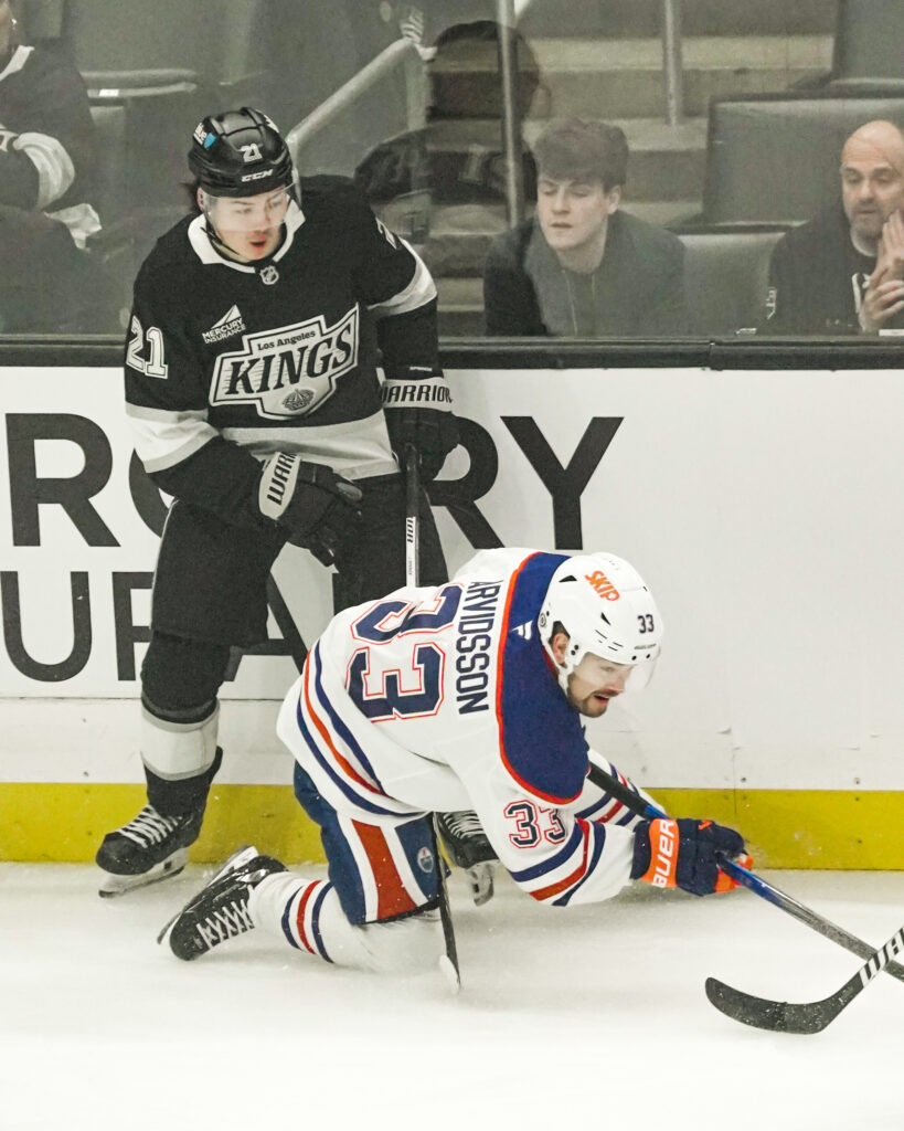 A hockey player from the Kings watches an Oilers player on the ice.