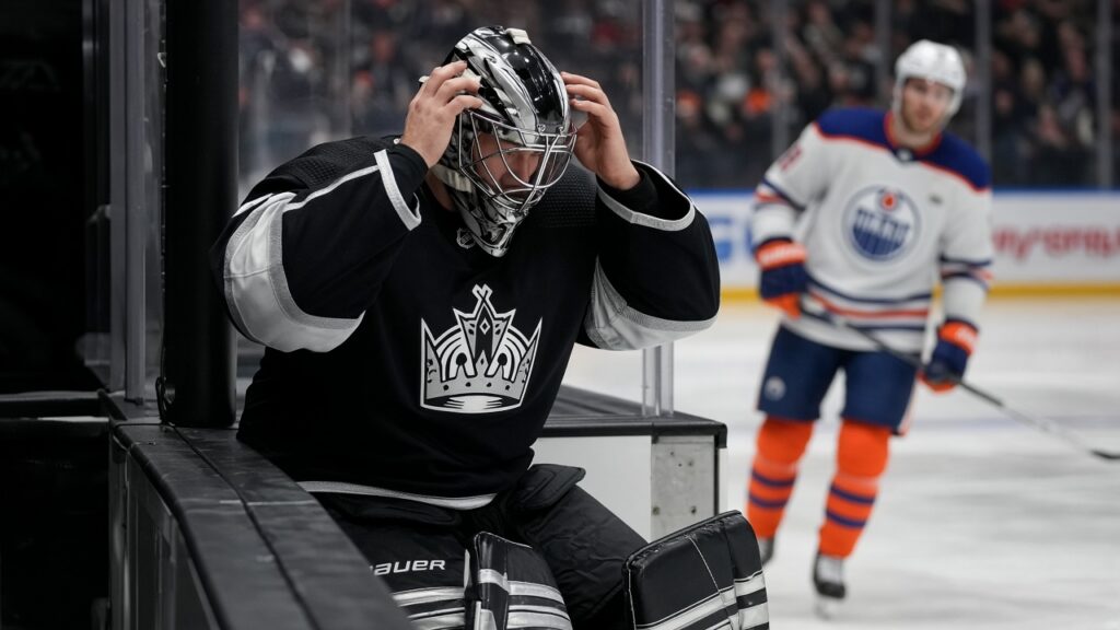 A Kings goalie in a black home jersey pulls on his mask at the bench as an Edmonton Oilers player skates past