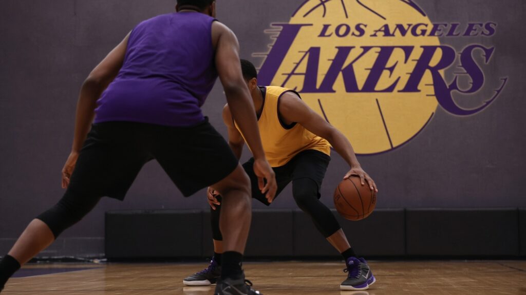 Lakers Training Camp Day 2: Redick Staggers Luka and Reaves as the Wings Draw a ‘Line of Confrontation’ A Lakers defender in a purple practice jersey crouches to guard a ball-handler in gold beneath the Los Angeles Lakers logo during training camp