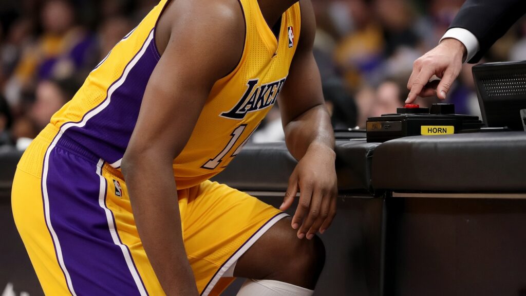 A Lakers player in a gold jersey crouches at the scorer's table waiting to check in as an official reaches for the horn